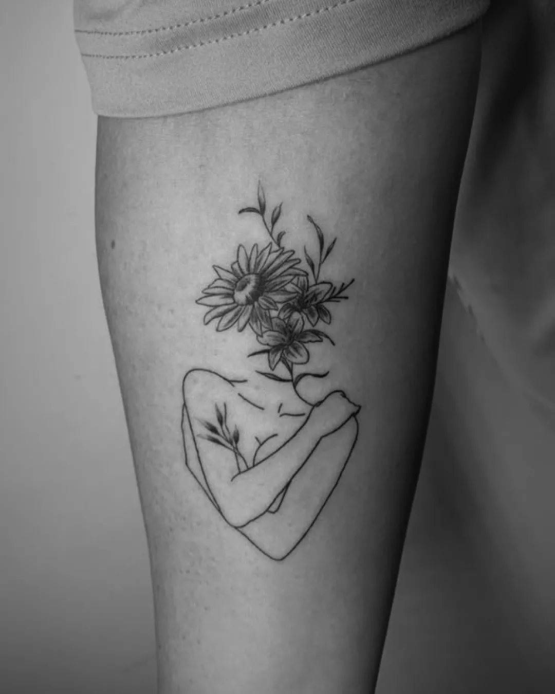 Fineline-Tattoo: Frauenkörper in feiner Linie, der sich selbst umarmt, mit Sonnenblume und Lilien anstelle des Kopfes